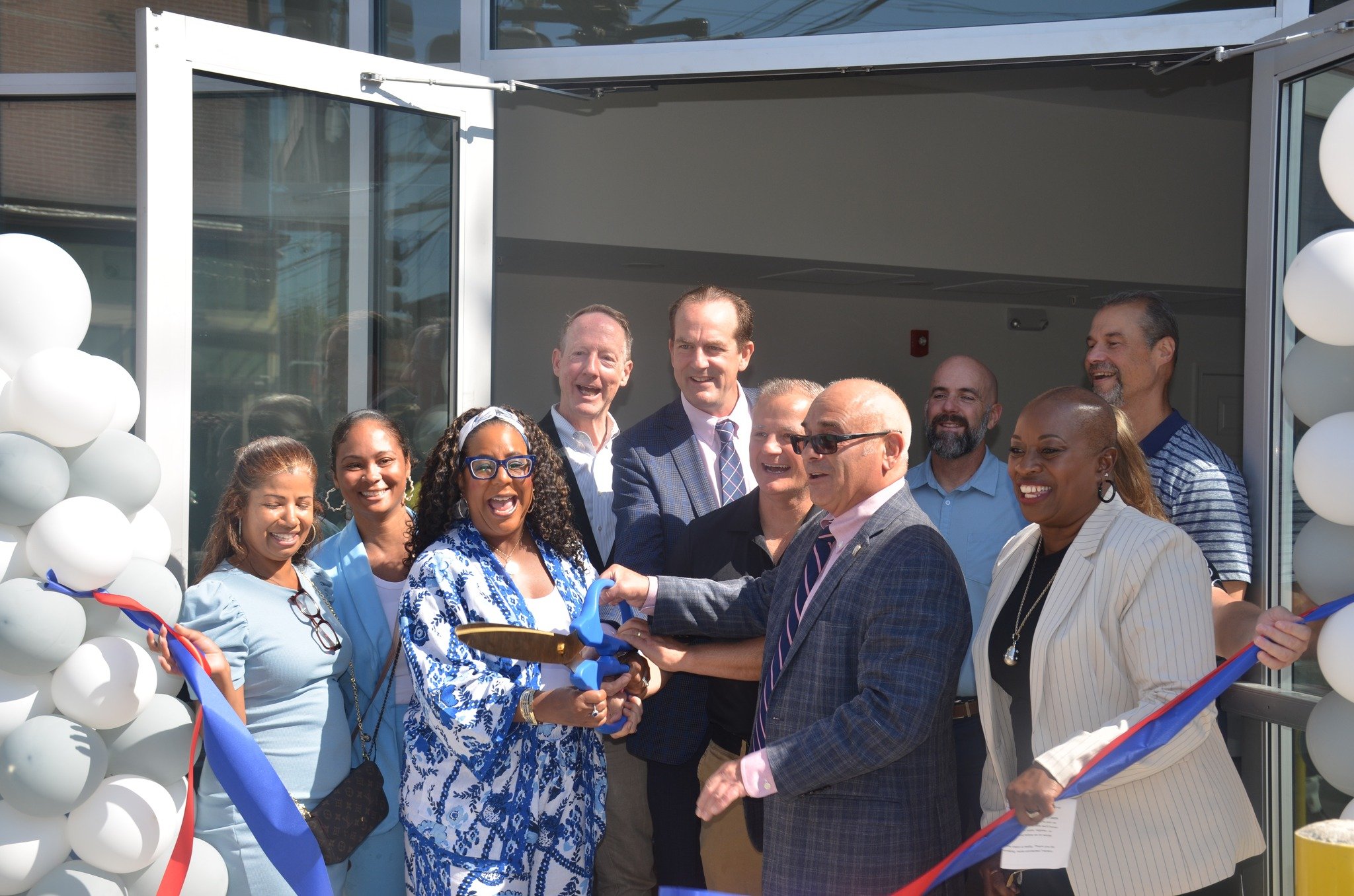 Group of city officials, community leaders, and developers smiling and cutting a ribbon with oversized scissors at the Clinton Lofts grand opening in Trenton, NJ.